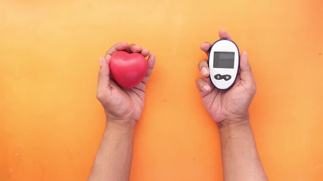 Close up of person's hands holding red heart and a glucometer on an orange background, symbolizing health, well-being, and medical care