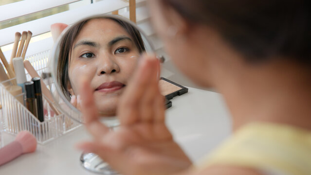 Young adult woman gen z black asia people relax get ready prepare clean makeup apply cream skin care make up getting dressed at home. Look at mirror camera self love anti aging health happy vanity.
