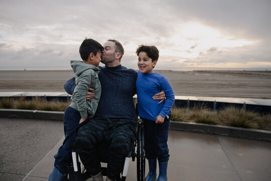 Father in Wheelchair With Sons Hugging and Smiling on the Beach