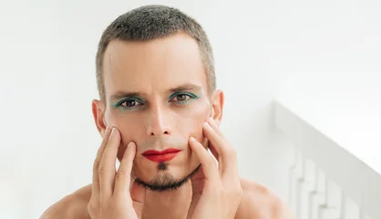 portrait of gay man with makeup and cosmetics on face. A handsome young transgender male with red lipstick on lips and mascara on eyes on a white background © alexkoral