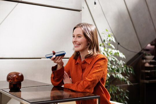 Smiling Woman Recording Voice Message on Phone at Caf&eacute; Table