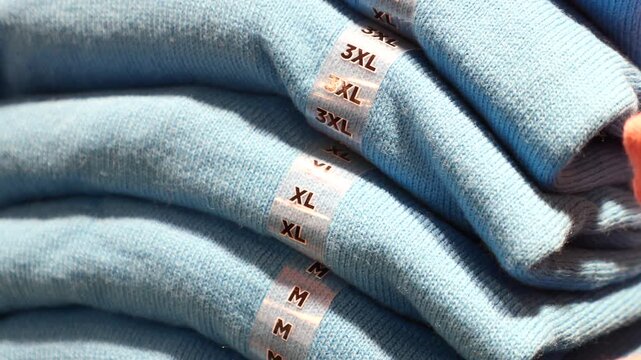 A close up view of neatly folded light blue knit sweaters with various sizes including 3XL, XL and M stacked in a retail store