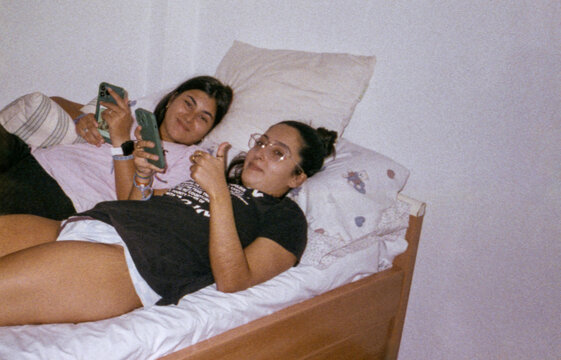 Two Young Women in Bed Looking at Camera with Phones in Hand