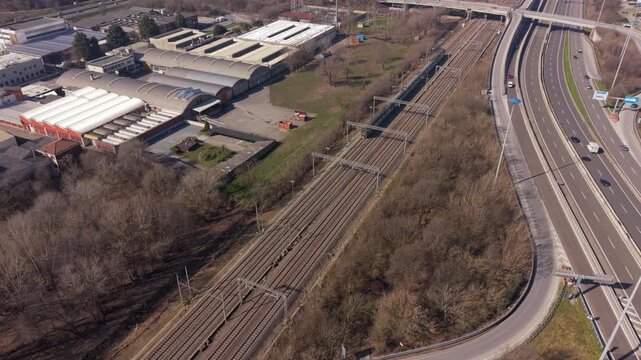 An expansive view of a complex railway network. Parallel tracks intertwine, leading towards industrial buildings and the intersecting presence of a highway.