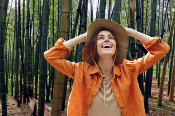Woman in hat stands amid tall bamboo forest, smiling and looking up with joy. Outdoor portrait, orange jacket, nature travel, peaceful green grove and happy candid expression. © SHOTPRIME STUDIO