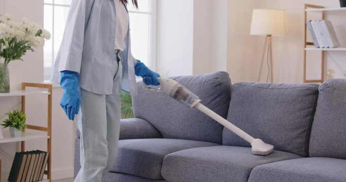 Woman cleaning couch cushions with vacuum cleaner extractor machine for dry clean at home. Worker of cleaning service in rubber gloves working with cordless electrical appliance for housekeeping