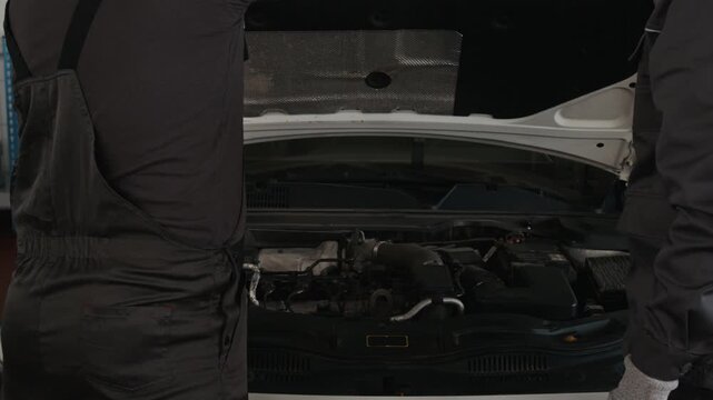 Medium rear shot of two professional Caucasian male mechanics opening car hood, propping with rod, then inspecting car engine and discussing problem with ignition or combustion systems