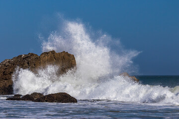 Obraz premium Giant wave breaking against large rock just offshore Malibu, California. Spray flying high in the air; blue sky in the distance. 
