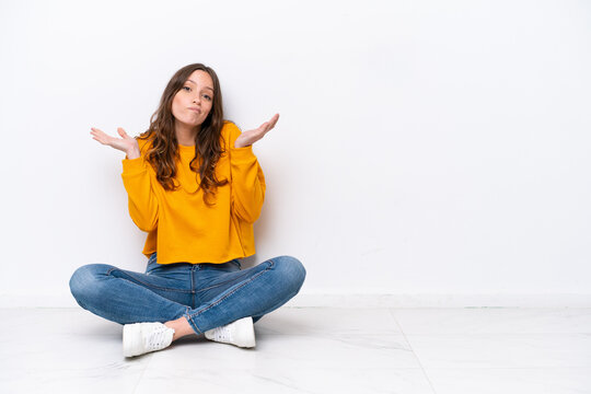 Young caucasian woman sitting on the floor isolated on white wall having doubts while raising hands
