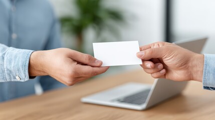 Obraz premium Two hands exchanging a blank business card over a wooden table with a laptop in the background and a green plant visible, ideal for networking or professional meetings