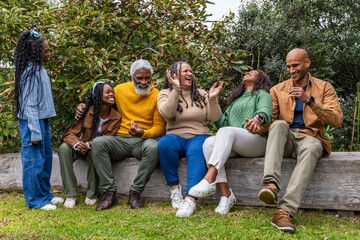 African American family in knit sweaters laughing, sitting on wooden log bench on lawn holding cup
