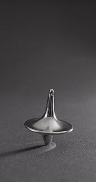 Metallic Spinning Top Falling Over. Linear footage of a shiny chrome top in steady rotation, progressively wobbling with increasing tilt until it fully topples on a matte gray surface.