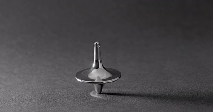 Metallic Spinning Top Falling Over. Linear footage of a shiny chrome top in steady rotation, progressively wobbling with increasing tilt until it fully topples on a matte gray surface.