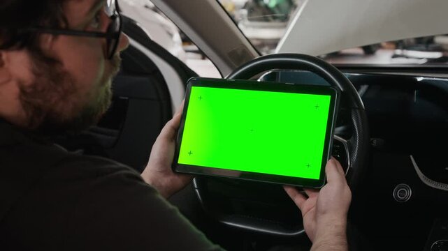 M shot of male car technician sitting at steering wheel in vehicle, holding digital tablet with green chromakey screen, performing OBD test to check engine performance