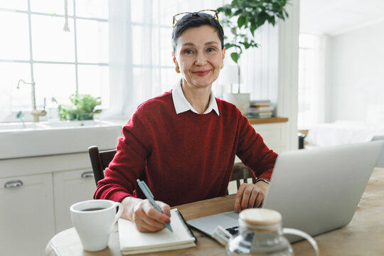 Indoor image of senior female ai trainer, establishing prompt testing working remotely from her cozy kitchen, sitting in front of laptop and looking at camera while writing in her copybook