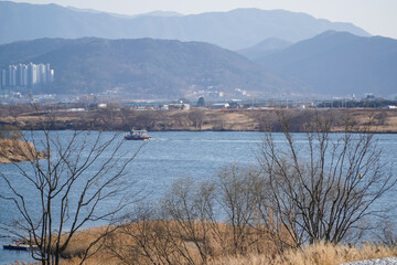 Obraz premium Boat Navigating a Wide River with Distant Mountains and Urban Buildings