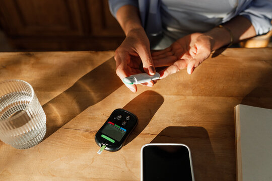 Picture of hands of unrecognizable woman doing finger prick test at home as morning routine to control level of blood sugar, suffering diabetes, sitting at sunlit table with measuring devices