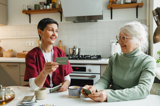 Games to eliminate and prevent dementia. Assisted living for old woman suffering memory loss, mature caregiver and her patient playing charades together at kitchen table holding cards with phrases