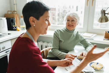 Selective focus on old smiling woman sitting at kitchen table near window and looking at her...