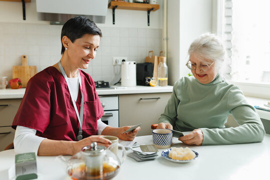 Dementia and alzheimer treatment games. Indoor picture of old woman with gray hair having fun playing cards with her service support volunteer, talking and drinking tea at daytime at kitchen