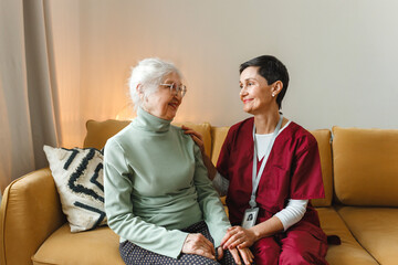 Woman medical worker in red uniform talking to her old female patient sitting on yellow couch,...