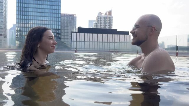 Bald man glasses kisses long-haired woman hot tub embrace. Urban high-rises frame romantic watery moment.