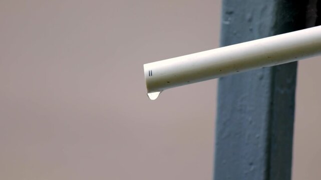 Single droplet suspends in mid-air, detaching from the tip of a white PVC pipe. The moment captures the quiet rhythm of dripping water, evoking a sense of stillness and subtle motion against a muted u