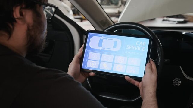 Medium over-shoulder shot of young Caucasian male mechanic with digital tablet sitting at steering wheel inside car, performing systems checkup using OBD testing software
