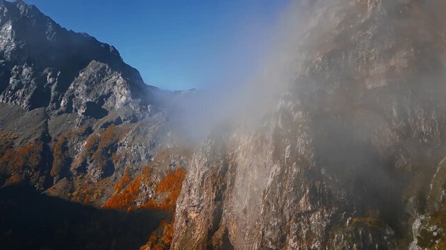 Aerial camera glides along steep limestone cliffs in Montenegro, low clouds stream, autumn beech patches in orange and gold cling to ledges under a clear sky.
