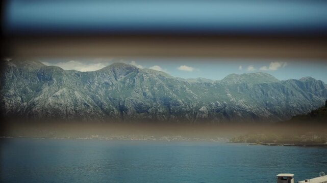Daytime view through three horizontal slats shows the Adriatic, karst cliffs, a small pier and villas, and clustered buildings across the bay in bright, calm light.