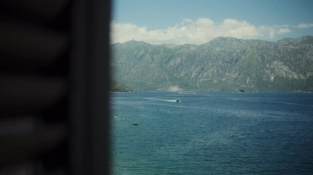 A framed window looks to Bay of Kotor, Montenegro, with shutters, gutter, small boats, buoys, and a speeding motorboat on calm water under soft midday clouds.