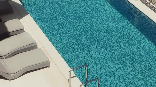 Narrow rectangular pool with mosaic tiles, stainless steel ladder, and three curved sun loungers on white deck by limestone wall in Montenegro under bright light