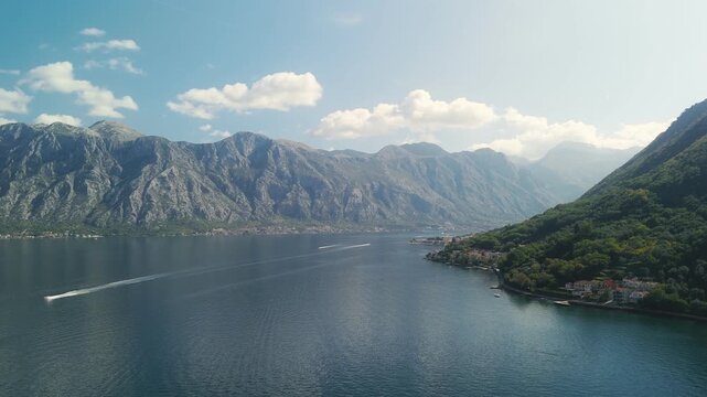 Aerial view of the Bay of Kotor, Montenegro. Speedboats leave white wakes as a ferry nears a coastal village. Karst limestone mountains rise under midday light and clouds.