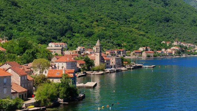 Aerial view in Montenegro shows a stone church bell tower, terracotta roofs, piers, and boats on a turquoise bay, with forested hillside, midday light, and wide composition.