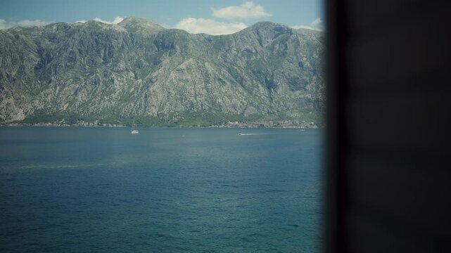 Daytime room to sea pano frames calm Adriatic water, karst mountains, coastal settlement, moored sailboat, and fast motorboat, Montenegro, clear sky, soft light.