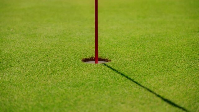 Tight view of a golf cup and red flagstick on a putting green in Montenegro by the Adriatic, a long shadow crosses the turf as the camera gently pushes in.