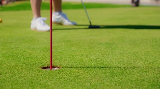 Low angle view shows a red flagstick, hole, and a golfer putting as the ball rolls into the cup on an Adriatic course in Montenegro in bright daylight.