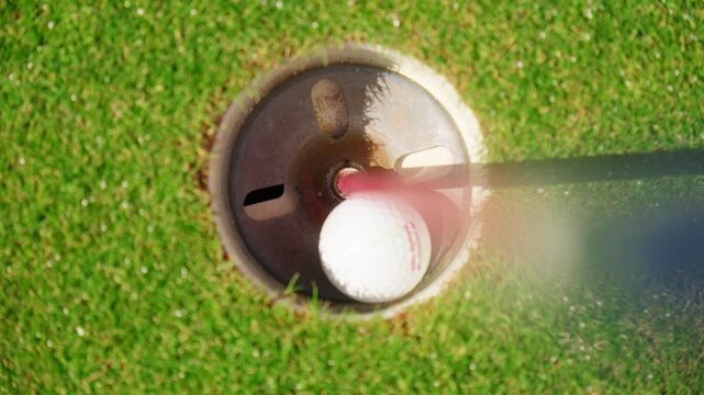 Top down view shows a red flagstick and white gulf ball drop into a metal lined cup on a putting green in Montenegro, with sharp shadow and motion blur in daylight.