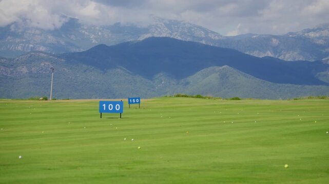 Wide driving range pan shows 100 and 150 markers on striped turf, many golf balls, and Montenegro mountain backdrop in daylight with vivid color and deep focus.