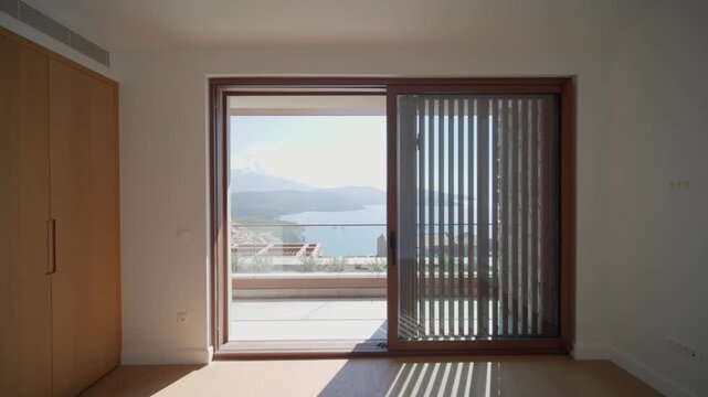 Symmetrical bedroom frames a sliding door and balcony. Vertical blind shifts, door moves, striped shadows pass on oak floor. Montenegro seaside and bay show.
