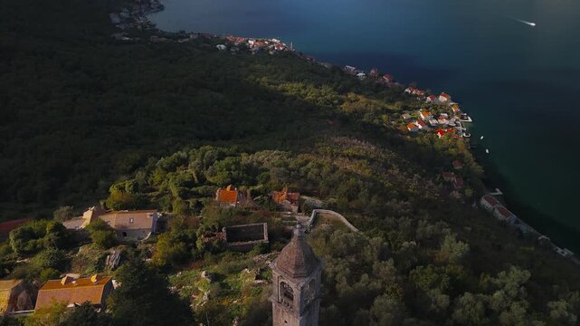 Aerial view shows stone church and bell tower on hillside in Montenegro, Our Lady of the Rocks, red roofs, olive groves, hamlets, motorboats, late afternoon light.