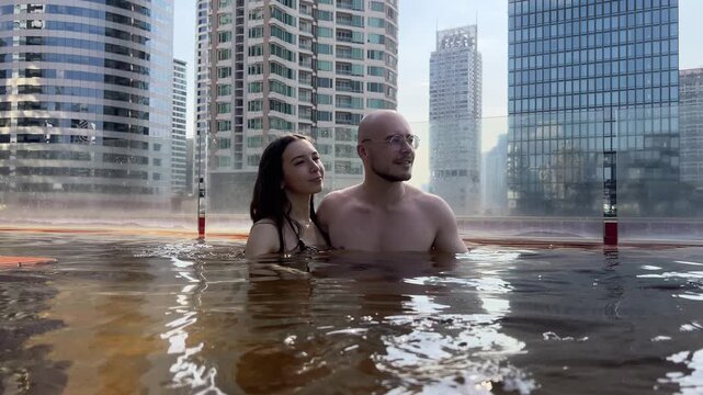 Shirtless man holds bikini woman waist wading hot tub. Urban towers frame romantic rooftop dip.
