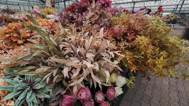 A dense and textured display of various high-quality artificial plants, featuring autumn leaves, large blossoms, and protea flowers in a garden center warehouse.