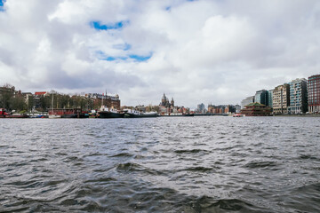 Naklejka premium Cityscape of amsterdam showing the amstel river, historic architecture along its banks, modern buildings, and various boats on a cloudy day, creating a panoramic view of urban life and travel
