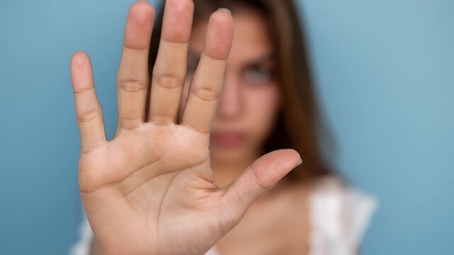 A young woman holds up her hand in a stop gesture against a blue background, expressing refusal and setting boundaries.