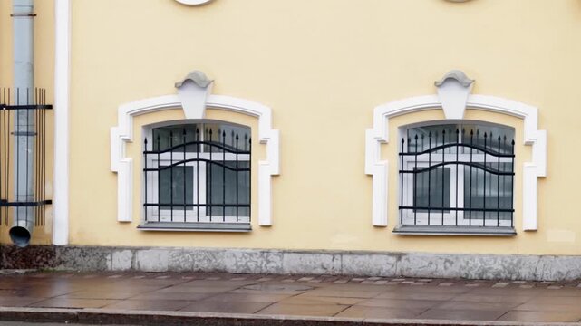 Pastel yellow building facade features two ornate window frames with decorative pediments and intricate black wrought iron grilles. Subdued colors and architectural details create nostalgic, serene mo