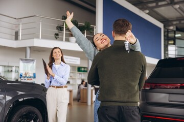 Obraz premium Happy couple hugging after buying new car at dealership, woman holding car key and saleswoman smiling
