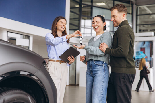 Salesperson handing over car keys to happy young couple purchasing new vehicle at dealership showroom