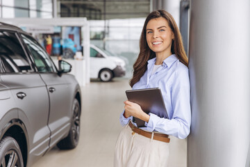 Saleswoman holding tablet smiling in car dealership showroom, advising and assisting clients with...