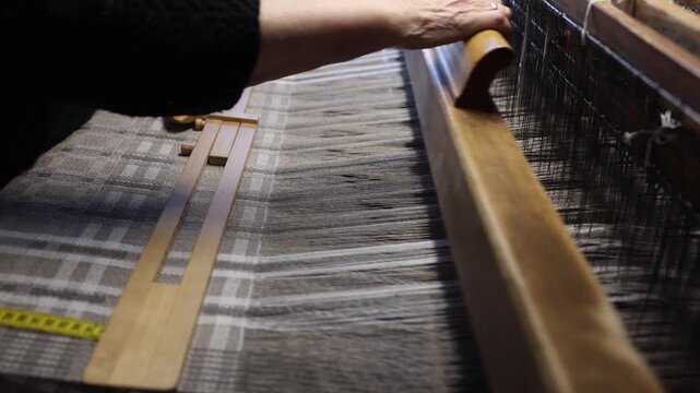 Close-up of hands operating a wooden loom, weaving patterned textile with visible threads and shuttle. Focus on craftsmanship and traditional textile production in a workshop setting